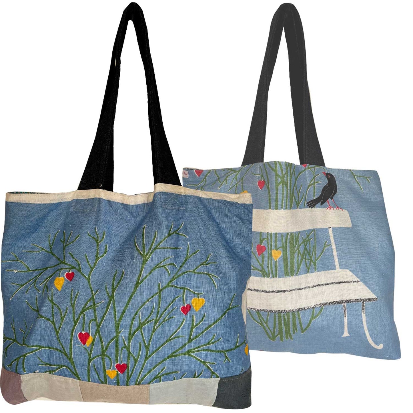 A light blue vintage teatowel tote bag with denim shoulder straps. a bench, a black bird, green grass, and small red and yellow hearts . Photographed on a white background.