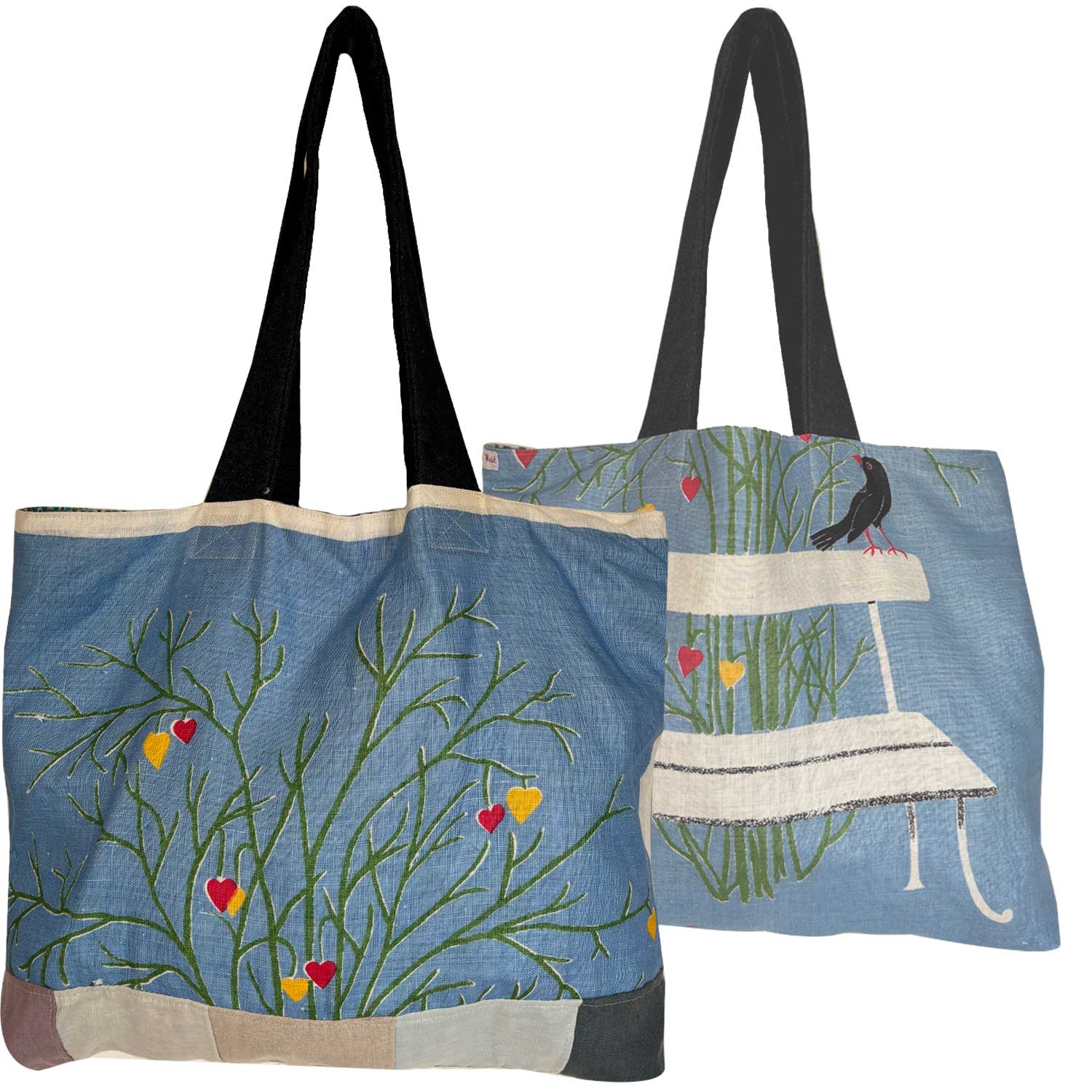 A light blue vintage teatowel tote bag with denim shoulder straps. a bench, a black bird, green grass, and small red and yellow hearts . Photographed on a white background.
