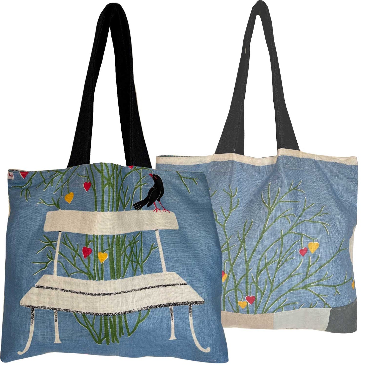 A light blue vintage teatowel tote bag with denim shoulder straps. a bench, a black bird, green grass, and small red and yellow hearts . Photographed on a white background.