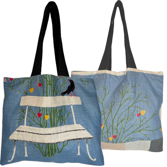 A light blue vintage teatowel tote bag with denim shoulder straps. a bench, a black bird, green grass, and small red and yellow hearts . Photographed on a white background.